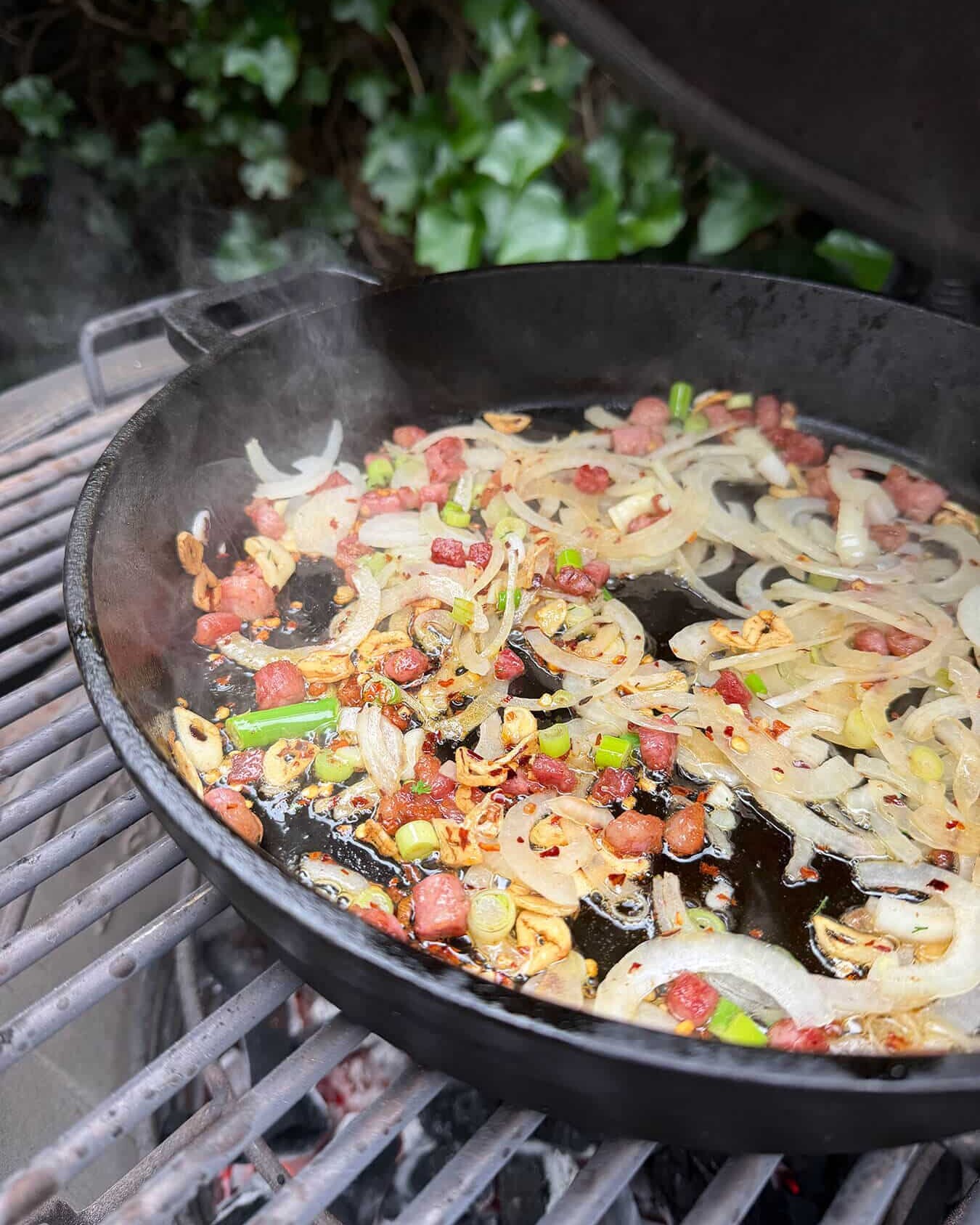 Bacon, uien en knoflook bakken in een gietijzeren pan op een grill. - Dutch BBQ Dude