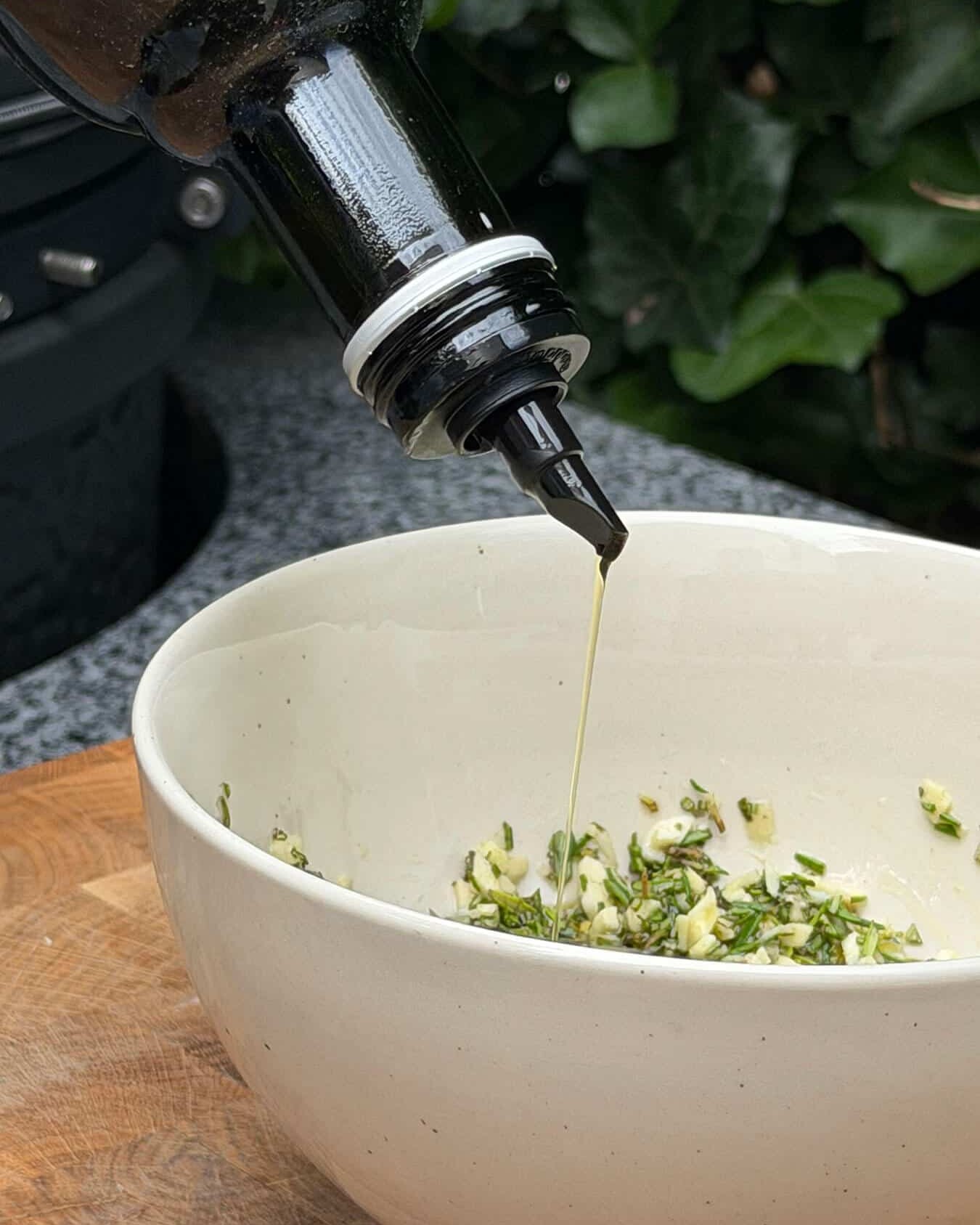 Olijfolie wordt toegevoegd aan een kom met knoflook en rozemarijn voor kip aan het spit. - Dutch BBQ Dude