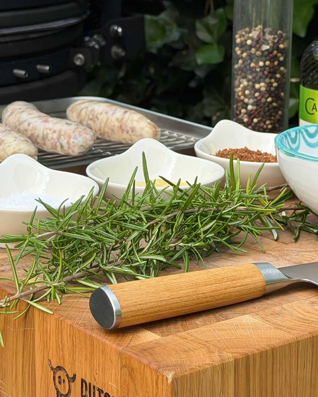 Varkensworstjes, rozemarijn, kruiden en een mes op een snijplank - Dutch BBQ Dude