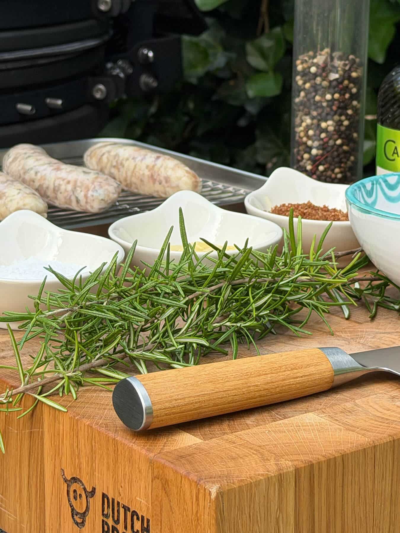 Varkensworstjes, rozemarijn, kruiden en een mes op een snijplank - Dutch BBQ Dude